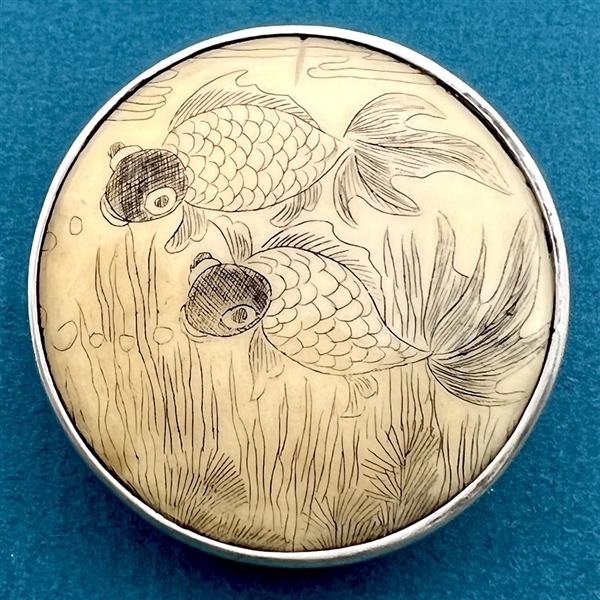 Incised, inked and painted natural material button of two fish swimming.